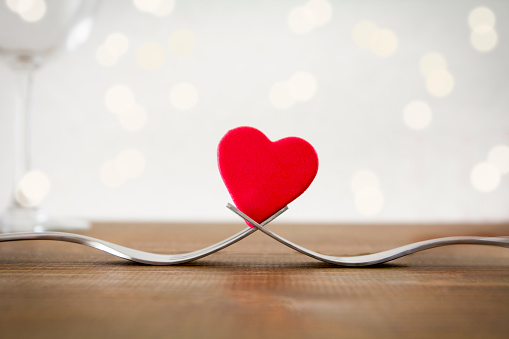 A red heart between two forks in a romantic dinner setting with defocused lights and wine glass.\nValentine's Day/ Romantic Dinner concept shot.