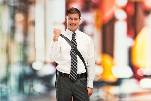 One person of with short hair caucasian young male businessman in front of defocused background who is outdoors wearing messenger bag who is successful and cheering and showing fist who is and doing fist pump and holding bag