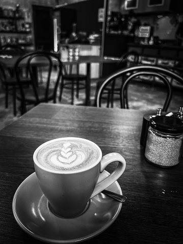 Vertical high angle closeup black and white photo of a cup of latte coffee on a wooden table in a cafe. Soft focus cafe background.