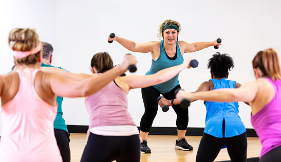 A fitness instructor leading an aerobics class. The instructor, a mature Hispanic woman in her 40s, is standing in front of the multiracial group of four women and one man, smiling and talking into a headset mic. She is squatting and lifting dumbbells with arms outstretched as everyone follows along in unison.