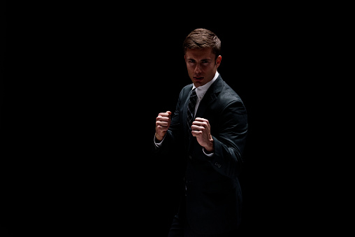 One person of aged 20-29 years old with brown hair caucasian male businessman in front of black background wearing businesswear who is serious and showing fist who is punching