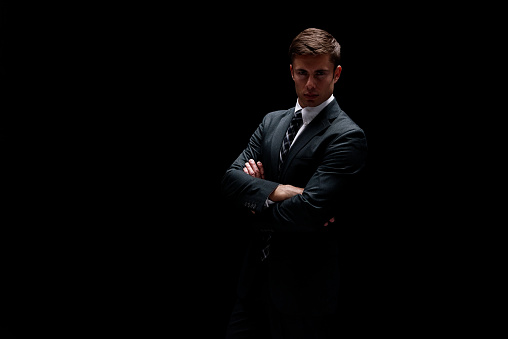 Waist up of aged 20-29 years old with brown hair caucasian male businessman in front of black background wearing businesswear who is in concentration with arms crossed
