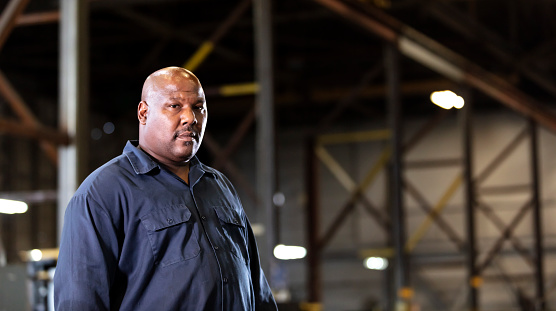 A mature African-American man with a large human build standing in a dimly lit manufacturing warehouse. He is looking away with a serious expression on his face. He is bald, with a mustache and hair stubble.