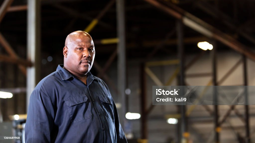 African-American man working in dark warehouse A mature African-American man with a large human build standing in a dimly lit manufacturing warehouse. He is looking away with a serious expression on his face. He is bald, with a mustache and hair stubble. African-American Ethnicity Stock Photo