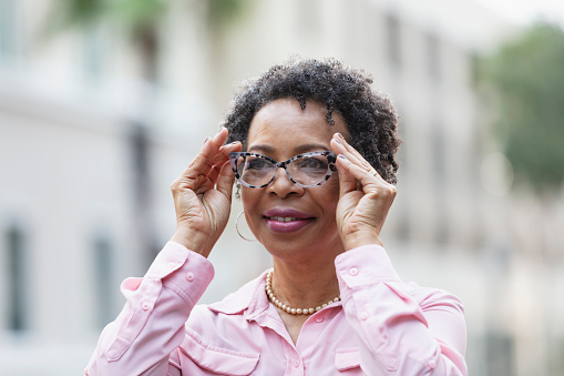 Headshot of a mature black woman walking outdoors in the city, putting on a pair of eyeglasses.