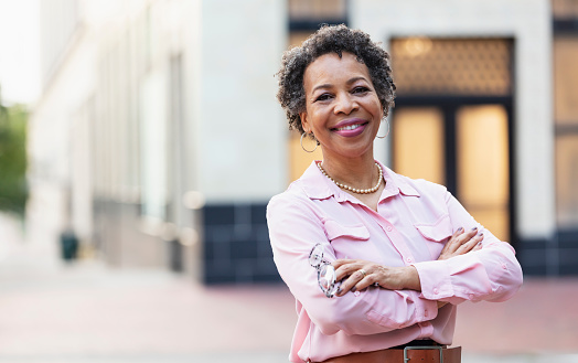 Portrait of a mature black woman standing outdoors in the city, smiling confidently at the camera, arms crossed.