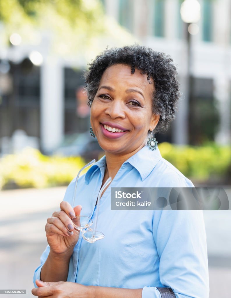 Mature black woman in city, smiling at camera A mature black woman in her 50s walking outdoors in the city, carrying a handbag, smiling at the camera. Mature Women Stock Photo