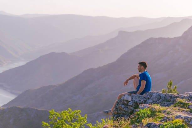 istock Adventurous man sitting on top of a mountain and enjoying the beautiful view, while looking downhill at the blue river and amazing mountain line. 1364684066