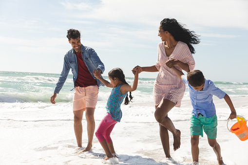 Happy multiracial parents holding hands of children while walking at beach enjoying sunny day. family, lifestyle and weekend.