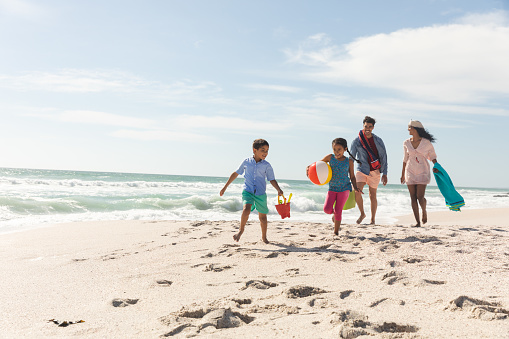 Padres multirraciales caminando detrás de los niños corriendo en la arena de la playa durante el día soleado photo