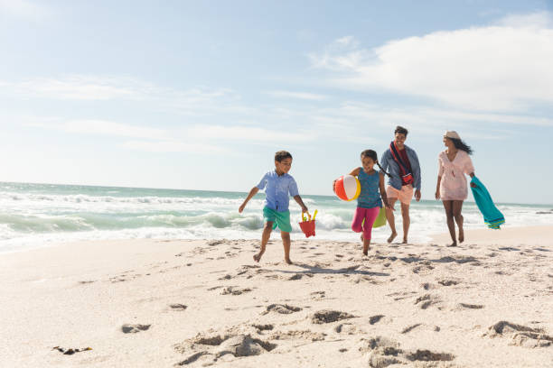 Multiracial parents walking behind children running on sand at beach during sunny day stock photo