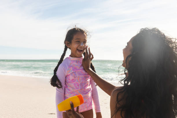 Cheerful biracial girl looking at mother applying sunblock lotion on her nose during sunny day stock photo