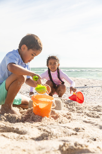 Biracial brother and sister collecting sand in buckets while playing at beach on sunny day. childhood and weekend.