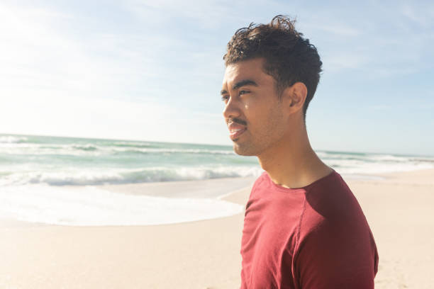 istock Biracial man looking away while standing at beach against sky on sunny day 1364697150