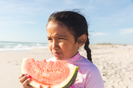 Cute biracial girl holding large fresh watermelon slice looking away at beach on sunny day. childhood and healthy food.