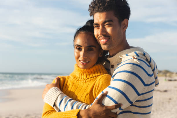istock Smiling biracial man embracing girlfriend while looking away at beach on sunny day 1364703022
