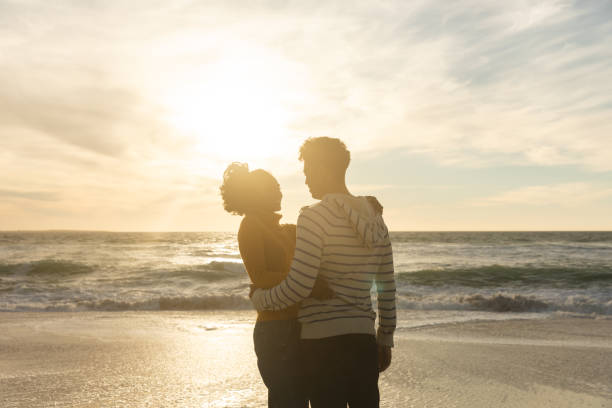 istock Romantic biracial couple embracing while standing at beach against sky during sunset 1364704589