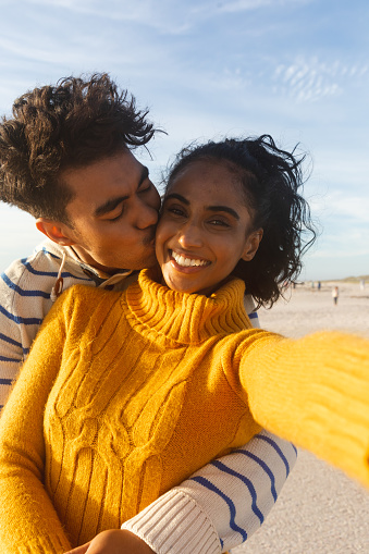Portrait of smiling biracial woman taking selfie of boyfriend hugging and kissing on cheek at beach. lifestyle, love and weekend.