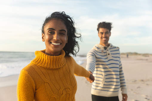 Portrait of smiling young biracial woman holding hand with boyfriend standing behind her at beach. lifestyle, love and weekend.