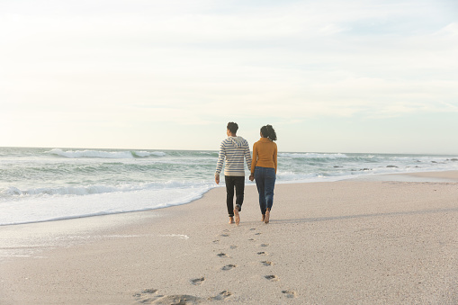 Full length rear view of biracial couple walking while leaving footprints on sand at beach. lifestyle, love and weekend.
