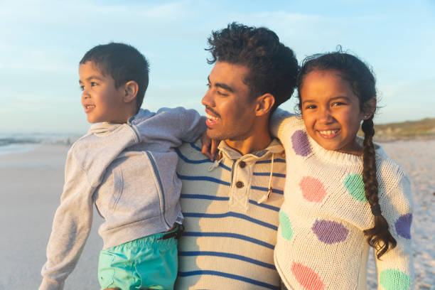 istock Happy biracial man carrying children while standing at beach against sky during sunset 1364711714