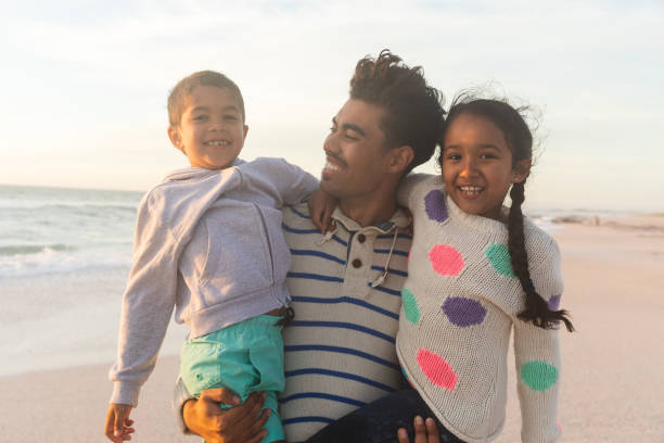 istock Happy biracial father carrying children while standing at beach against sky during sunset 1364711904