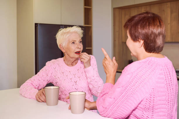 dwie stylowe seniorki w różowych swetrach siedzące z kubkami w nowoczesnej kuchni plotkują. - domestic kitchen family couple coffee zdjęcia i obrazy z banku zdjęć