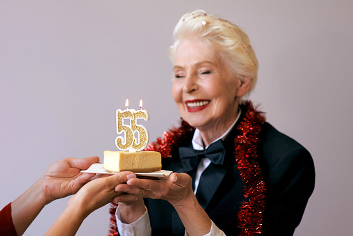 senior stylish woman in tuxedo blowing candle fifty five on her birthday cake. Lifestyle, positive, fashion, style concept