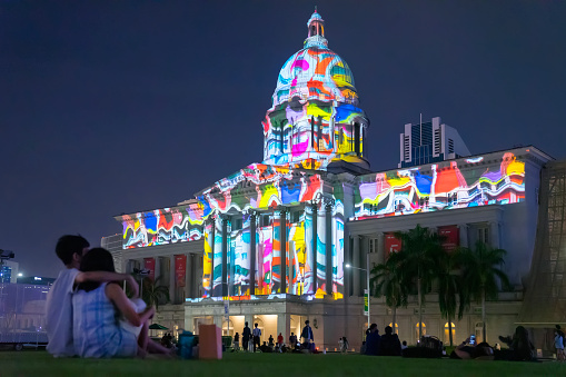 Singapore, Singapore - January 14, 2022: A couple watches as a colourful pattern is projected on the facade of the National Gallery of Singapore at night, as part of the 'Light To Night' segment of Singapore Art Week 2022.