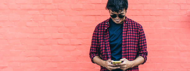 istock Horizontal banner with smiling asian young guy using mobile phone outdoor against a red wall - Asian social influencer having fun with new trends smartphone apps - youth millennial people lifestyle 1364727619