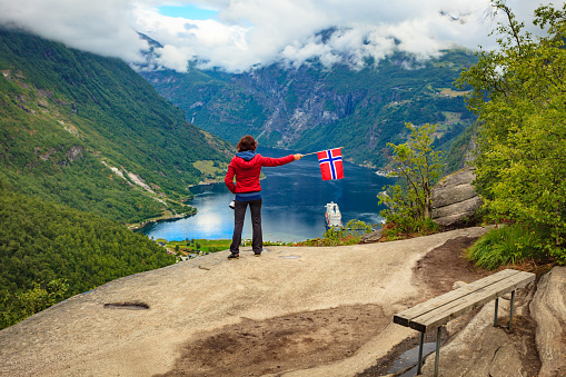Female tourist enjoying scenic view over fjord Geirangerfjorden from Flydalsjuvet viewpoint, holding norwegian flag. Cruising vacation and travel.