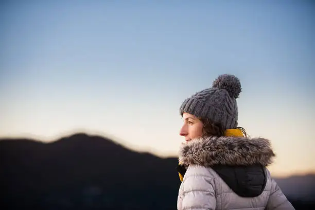 Side view of a beautiful young woman standing against a mountain range at dusk