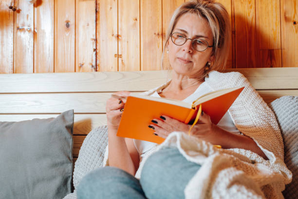 Close-up caucasian adult woman wearing glasses freelancer or student with diary sitting on bed in bedroom, indoors. Middle aged woman taking notes, reading something in notebook stock photo