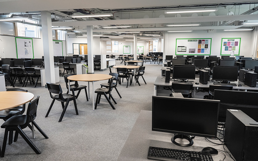 Wide angle view of unoccupied open plan room with desktop PC stations for individual work and round tables with chairs for student group discussions.