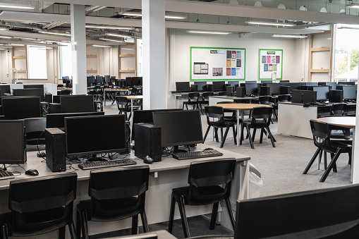 Unoccupied tables and chairs for collaborative educational work between rows of flexible workstations with desktop PC, keyboard, and mouse at each place.