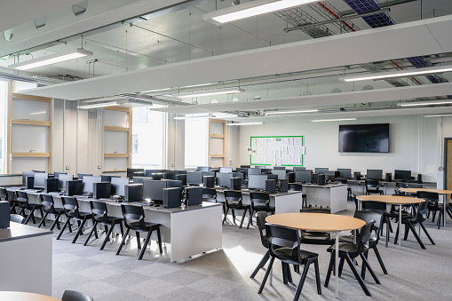 Wide angle view of unoccupied education environment with neatly arranged computer workstations and round tables for student group work.