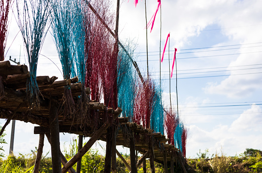 A low, close-up view of a bridge of eucalyptus logs decorated with brightly colored twigs, stretching along a cloudy road as a backdrop in the agricultural countryside of Thailand.