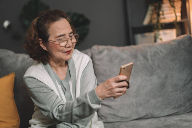 istock attractive Senior Asian woman Smiling while texting or chatting on smartphone at home. Old females or Grandmother holding mobile phone and playing social media or online podcast in the living room. Senior savvy with technology concepts 1364741496