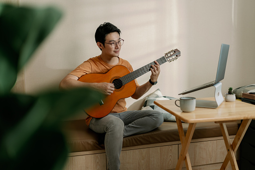 Asian male playing guitar Teleconferencing with music with his friend via video call on internet. Man having fun playing classical guitar. Musician man teaching online guitar lesson with his student on laptop computer. Online music class