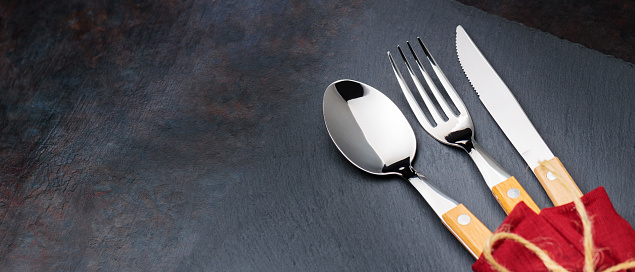 Cutlery with a burgundy napkin on a dark background. Spoon, fork and knife on a slate board. Table setting concept. Copy space. Top view