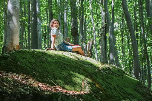 a teenage girl sits on a large mossy stone in the forest in the summer, in the Carpathians