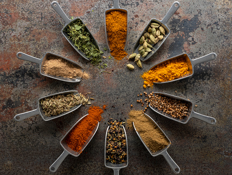 Many colorful, organic, dried, vibrant Indian food, ingredient spices in small aluminum metal spice scoops are arranged in a circle on an old, weathered, rusted metal plate background. Taken overhead, directly above the subject.