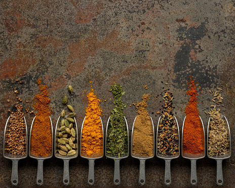Many colorful, organic, dried, vibrant Indian food spices in metal measuring dried food scoops on an old weathered abstract metal background.