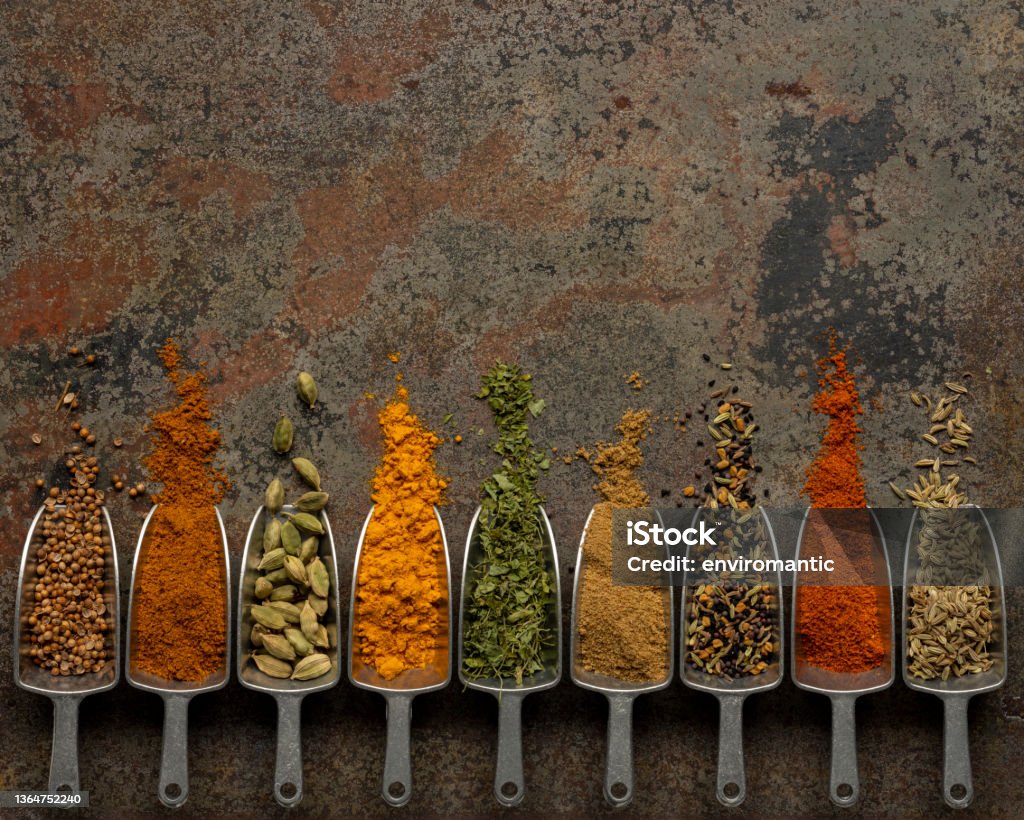 Many colorful, organic, dried, vibrant Indian food spices in metal measuring dried food scoops on an old weathered abstract metal background. Many colorful, organic, dried, vibrant Indian food, ingredient spices in small aluminum metal spice scoops are arranged in a line on an old, weathered, rusted metal plate background. Taken overhead, directly above the subject. Spice Stock Photo