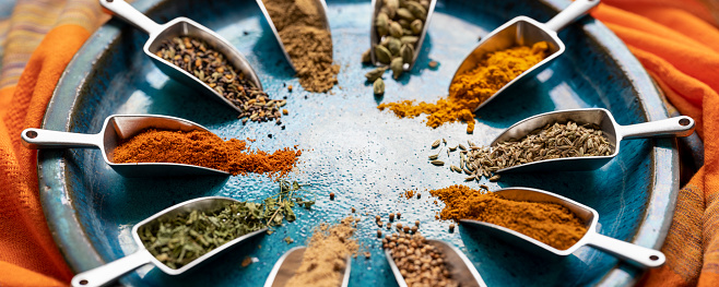 Many colorful, organic, dried, vibrant Indian food, ingredient spices in small aluminum metal spice scoops are arranged in a circle on an old blue-colored ceramic plate background, sitting on a vibrant, colorful contrasting textile. Very shallow depth of field, good copy space in the center of the image.