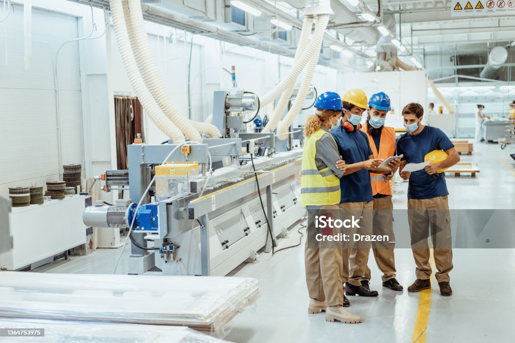 Employees in factory wearing face masks during meeting Engineers, architects and blue collar workers in protective clothing working in factory with modern machinery Manufacturing Stock Photo