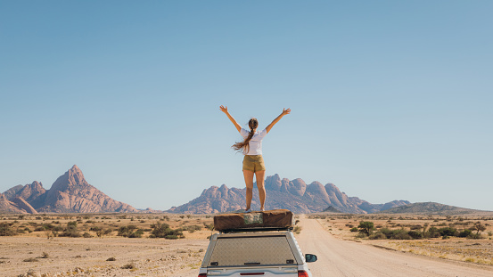 Young woman with long hair feeling freedom staying on the roof tent of the 4X4 camper truck, contemplating the road trip along the dramatic mountain landscape in Spitzkoppe