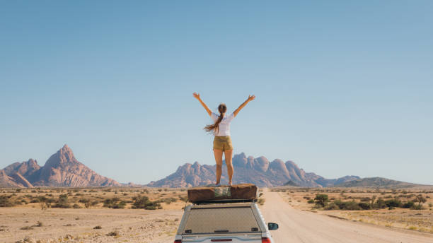 Female traveler staying on top of the camper car enjoying the view of scenic landscape in Namibia Young woman with long hair feeling freedom staying on the roof tent of the 4X4 camper truck, contemplating the road trip along the dramatic mountain landscape in Spitzkoppe southern africa stock pictures, royalty-free photos & images