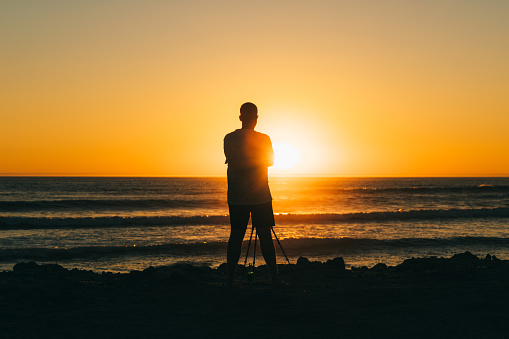 Male traveler staying at the beach taking photos of the beautiful colorful sunset by camera and tripod at the Skeleton Coast, Southern Africa