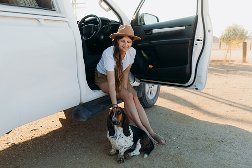Young happy woman in hat and the Basset Hound dog parking by the Atlantic Ocean with 4X4 camper truck, admiring the sunny sunset in Skeleton Coast National park